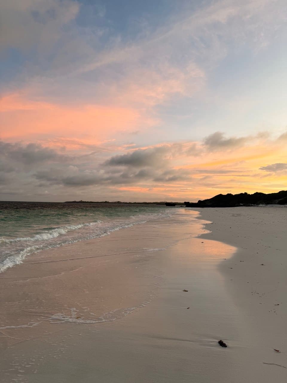 The Watamu beach at sunset: small waves washing over wet sand under a pink and orange sky, with the rocky headland dark in the distance.