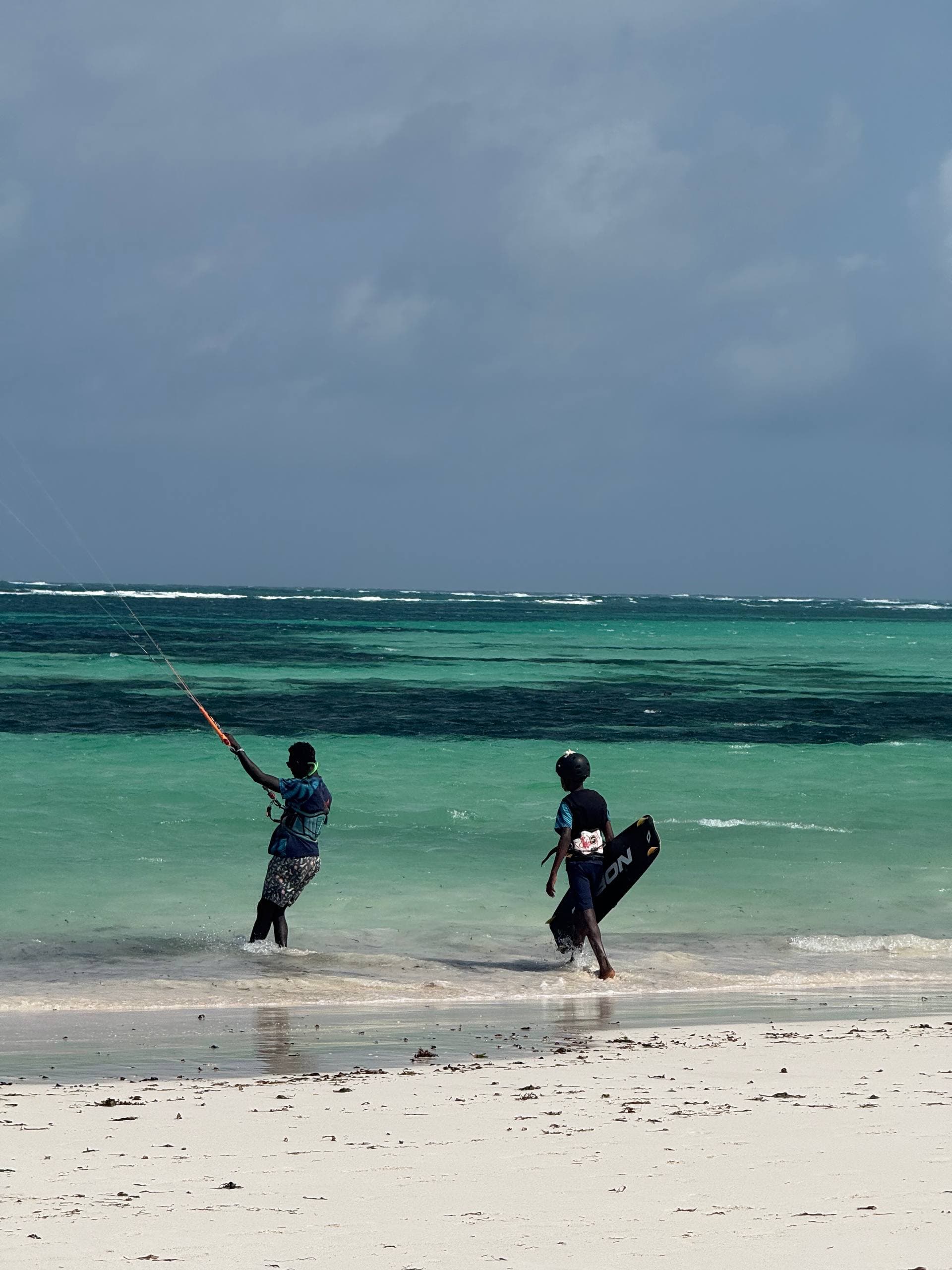 A Wind Family instructor steers a kite beside a young rider carrying a board, walking out of the turquoise shallows onto the white sand at Watamu