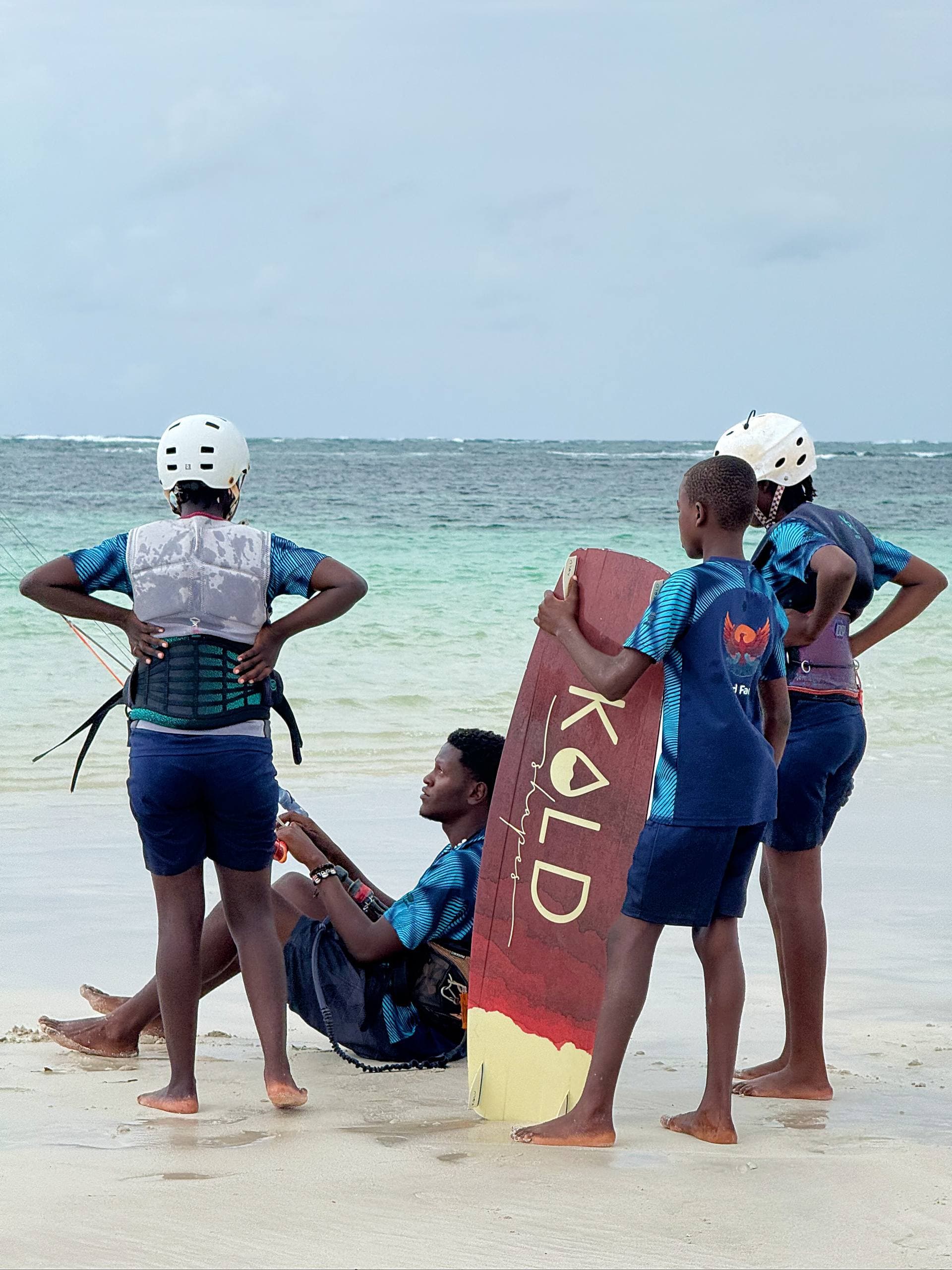 A Wind Family instructor sits on the sand holding a board while three young riders in helmets and impact vests stand around him at the water's edge in Watamu