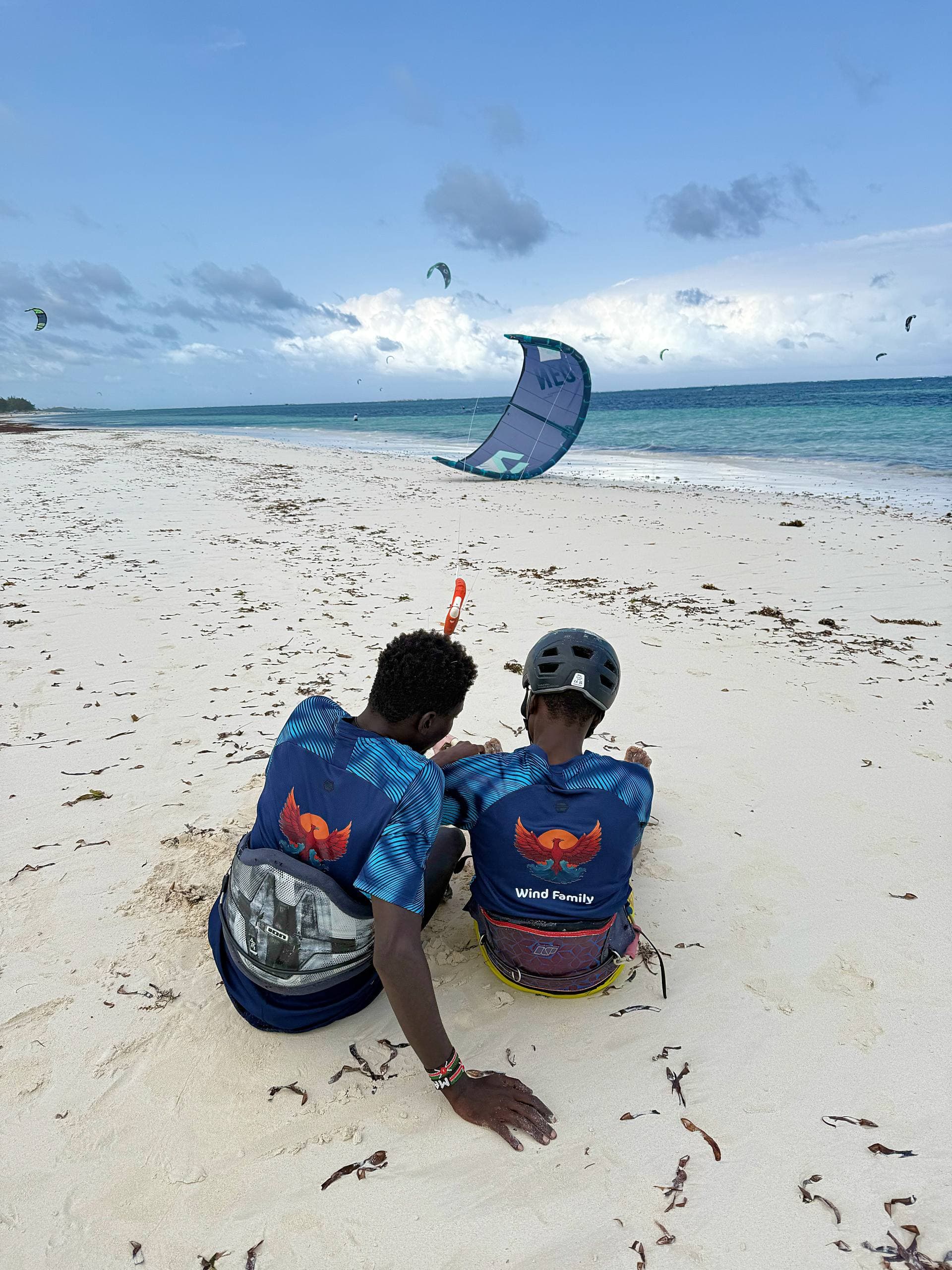 Two young riders in Wind Family jerseys sit on the sand watching a kite fly over the Watamu shoreline