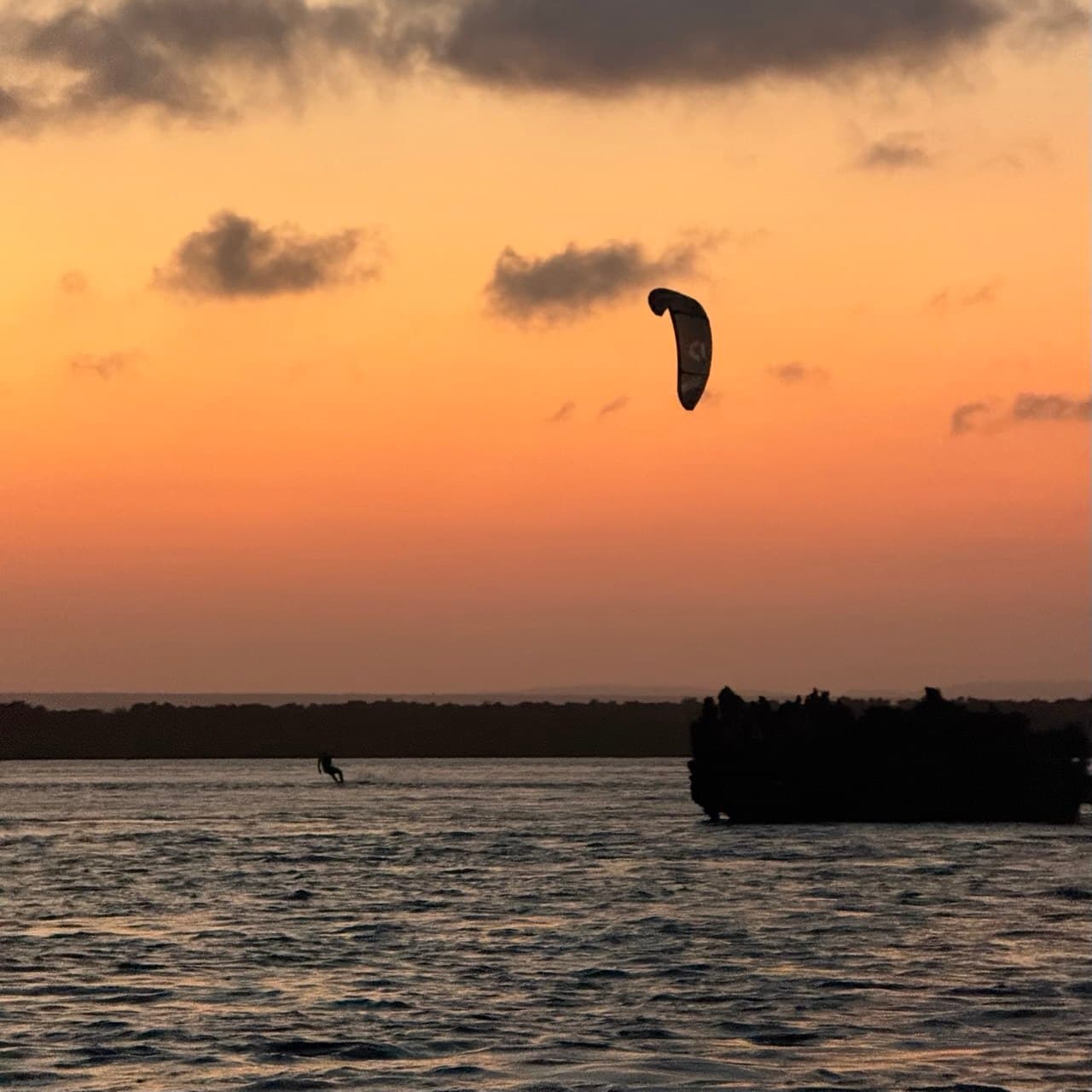 A kitesurfer rides across the Watamu lagoon at sunset, silhouetted against an orange sky with a boat in the distance.